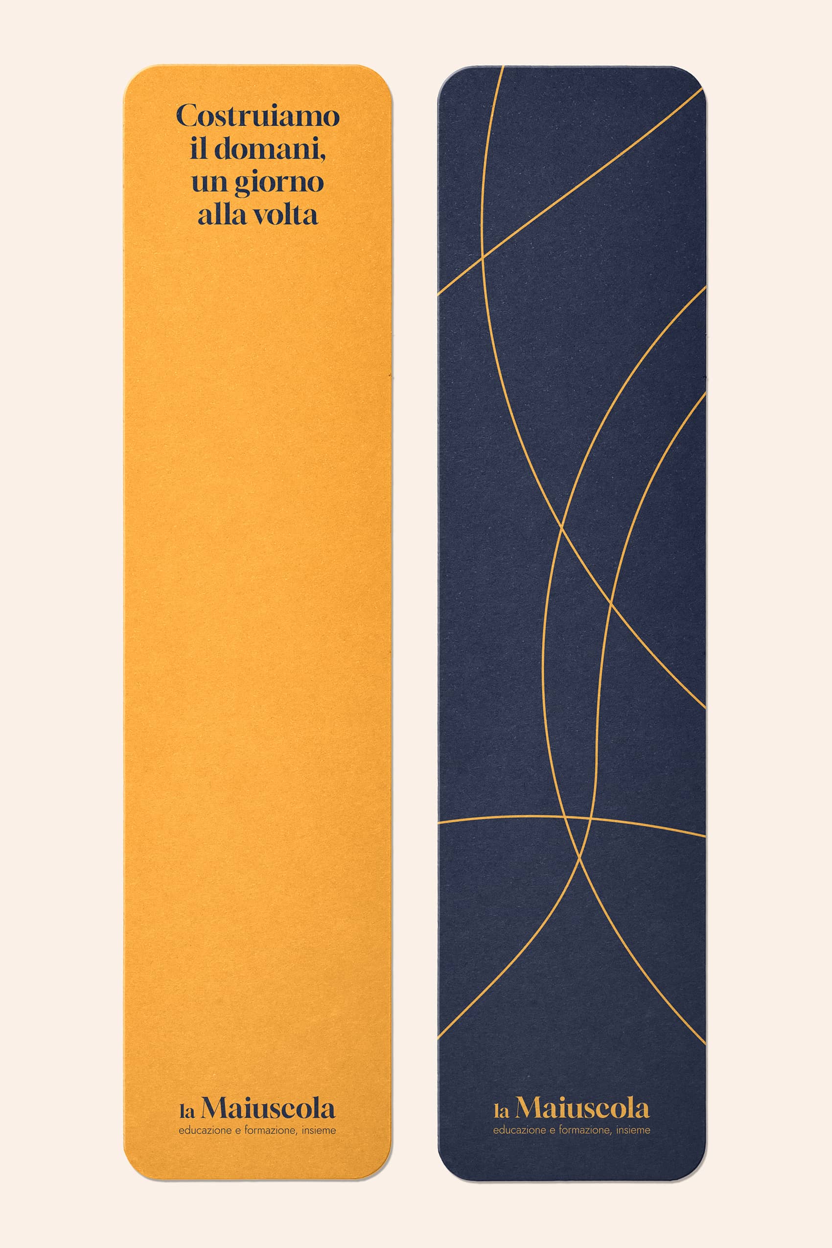 Two bookmarks side by side, one orange with Italian text 'Costruiamo il domani, un giorno alla volta' and 'la Maiuscola educazione e formazione, insieme' at the bottom, the other dark blue with abstract orange curved lines and the same text at the bottom.