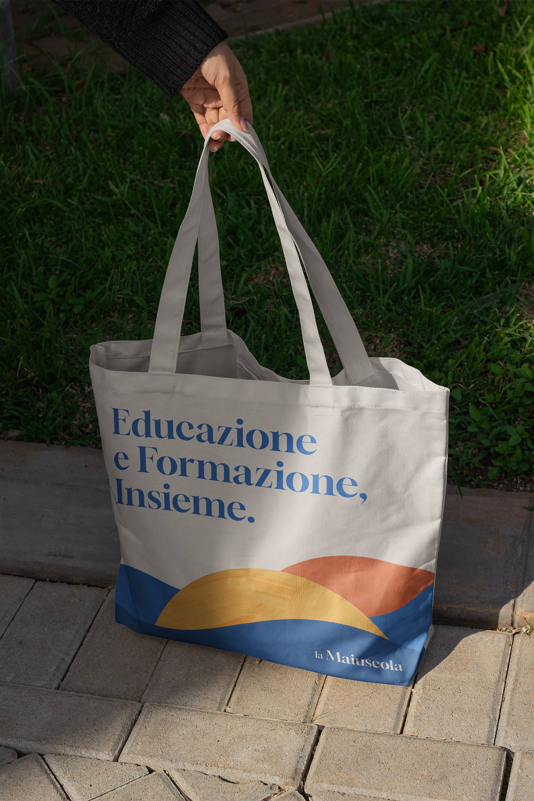 Hand holding a beige tote bag with Italian text 'Educazione e Formazione, Insieme.' and colorful abstract shapes at the bottom.