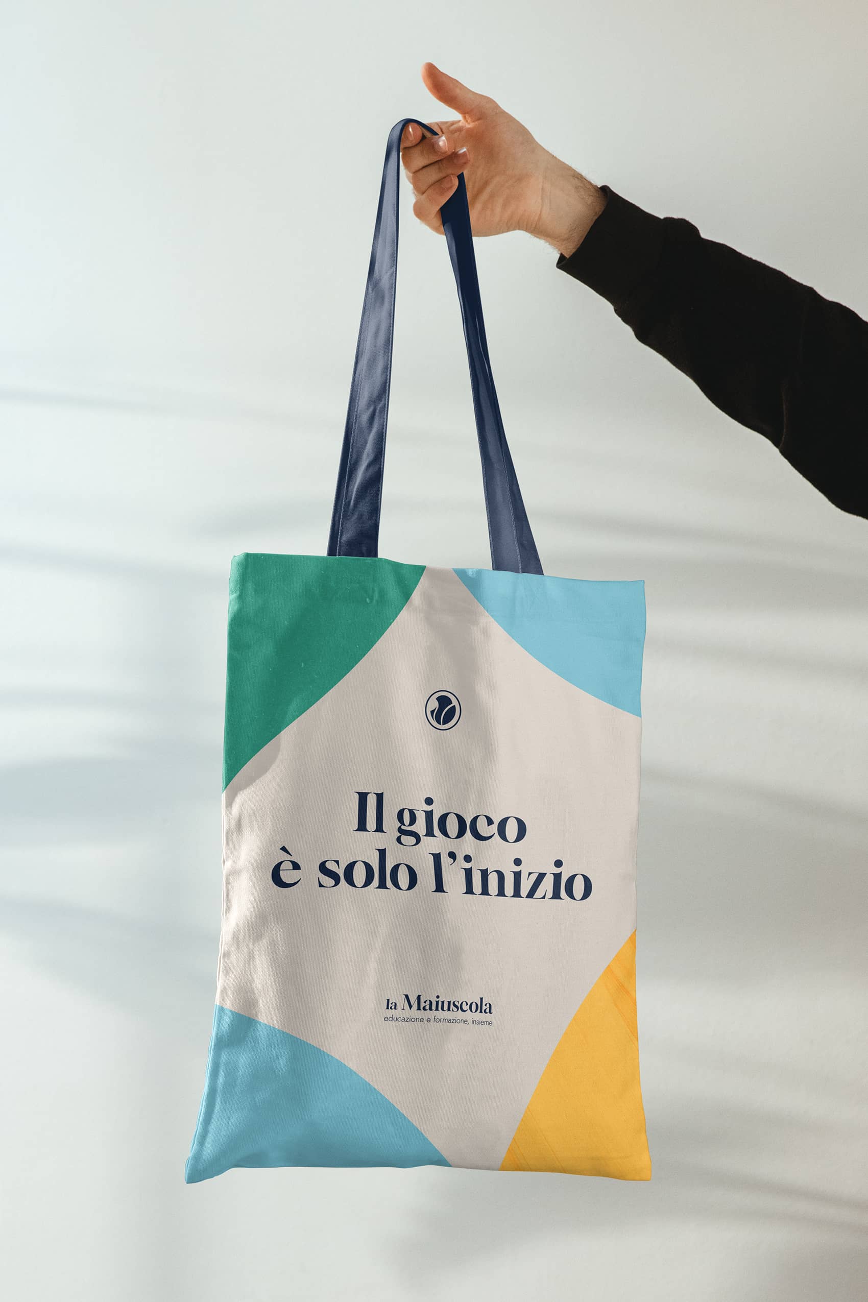 Hand holding a tote bag with blue handles and colorful geometric patterns, displaying Italian text 'Il gioco è solo l'inizio' and 'la Maiuscola' branding.