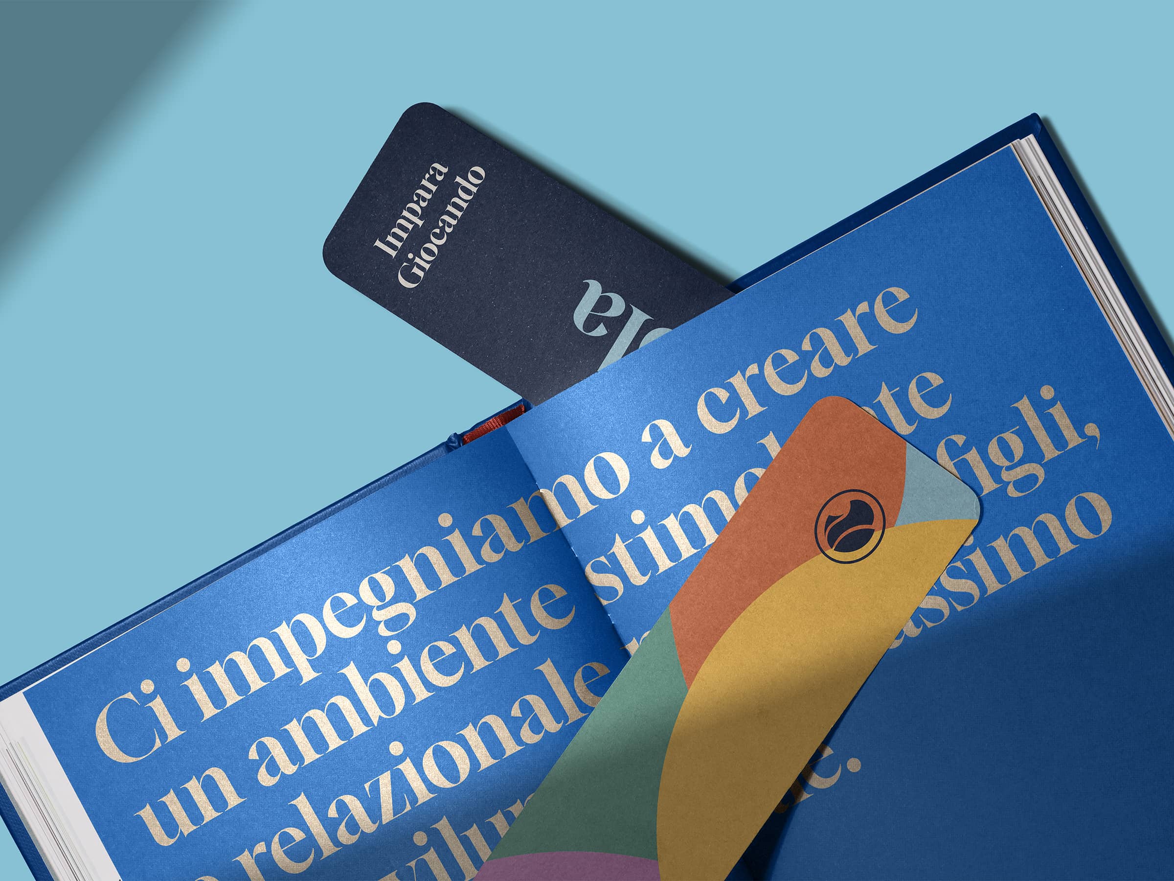 Open blue book with large beige Italian text and two colorful bookmarks, one navy with 'Impara Giocando' and one multicolored with a black logo.