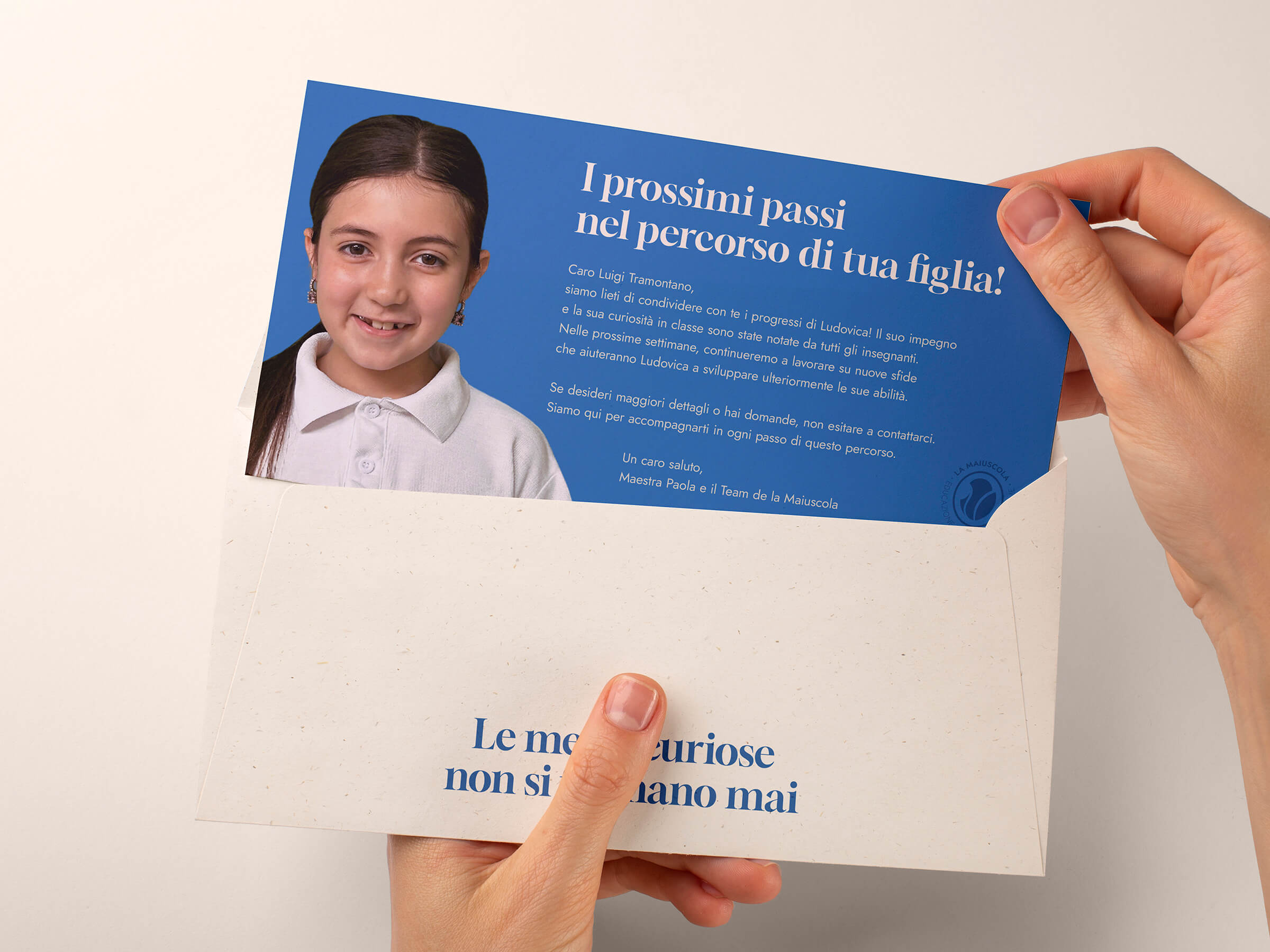 Hands holding an envelope with a blue card showing a young girl smiling and Italian text about her progress at school.