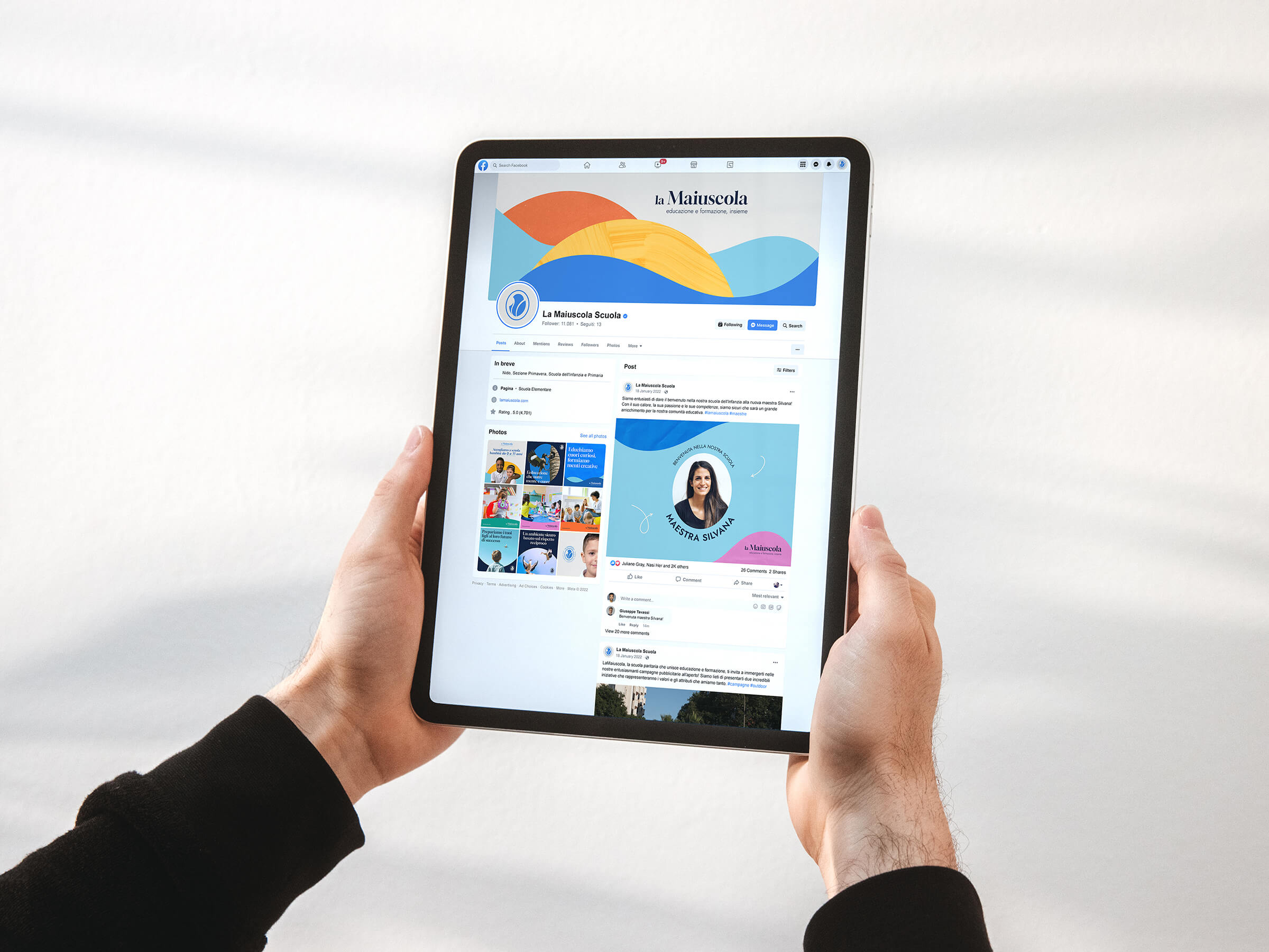 Hands holding a tablet displaying the La Maiuscola Scuola Facebook page with colorful banner and a post featuring a teacher's photo.